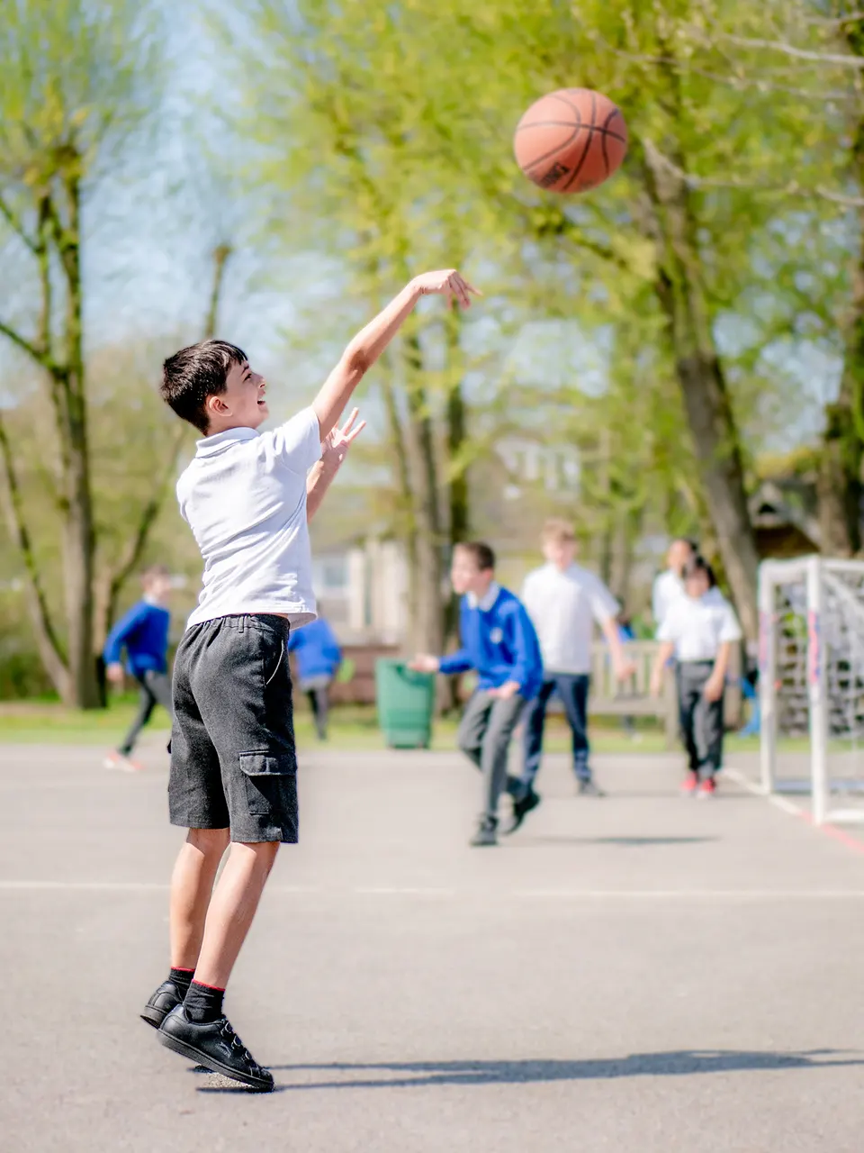 Boy shooting a basketball in playground with children behind playing football